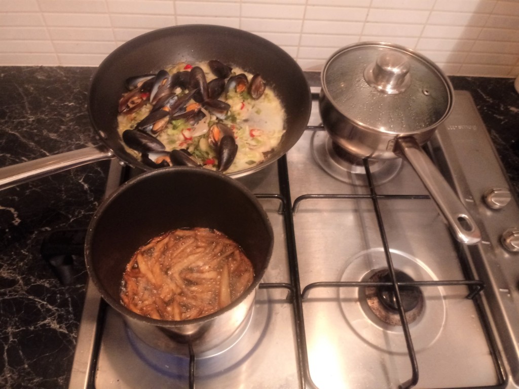 A pan of delicious mussels and colourful vegetables, some deep-frying potato chips, and a metal pan of recently-boiled water 