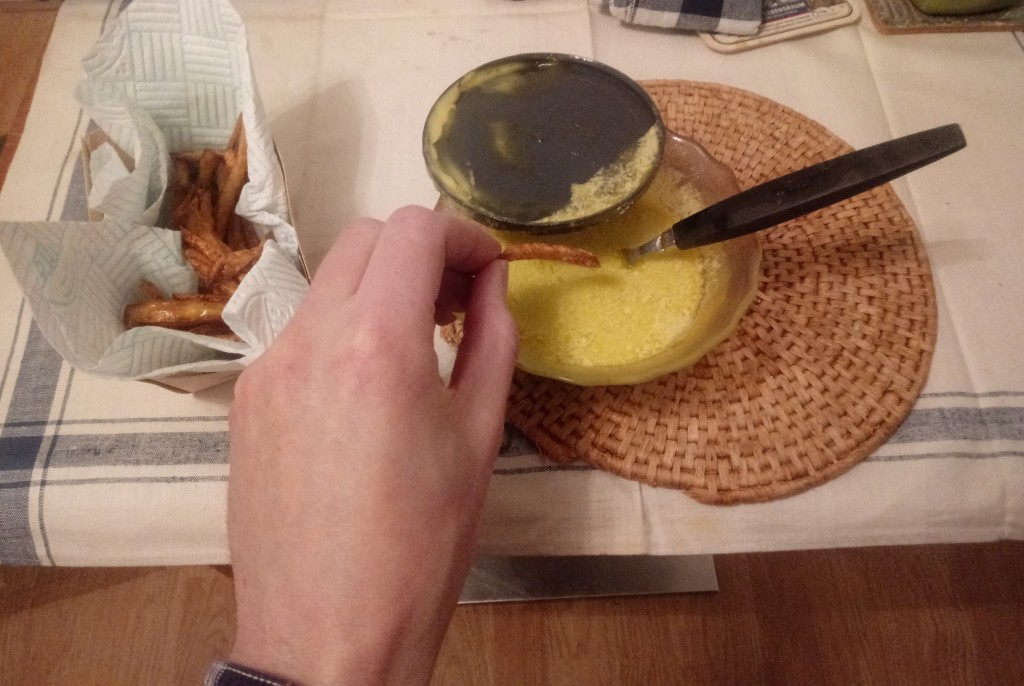 A white man's left hand dips a small, russet-coloured fry into a glass bowl of homemade yellow mayonnaise with a black spoon in it