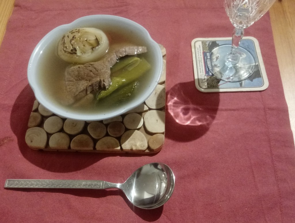 A blue ceramic bowl of Austrian Tafelspitz on a wooden place mat, red tablecloth and woodern table with a metal spoon and a wineglass on a white stork placemat